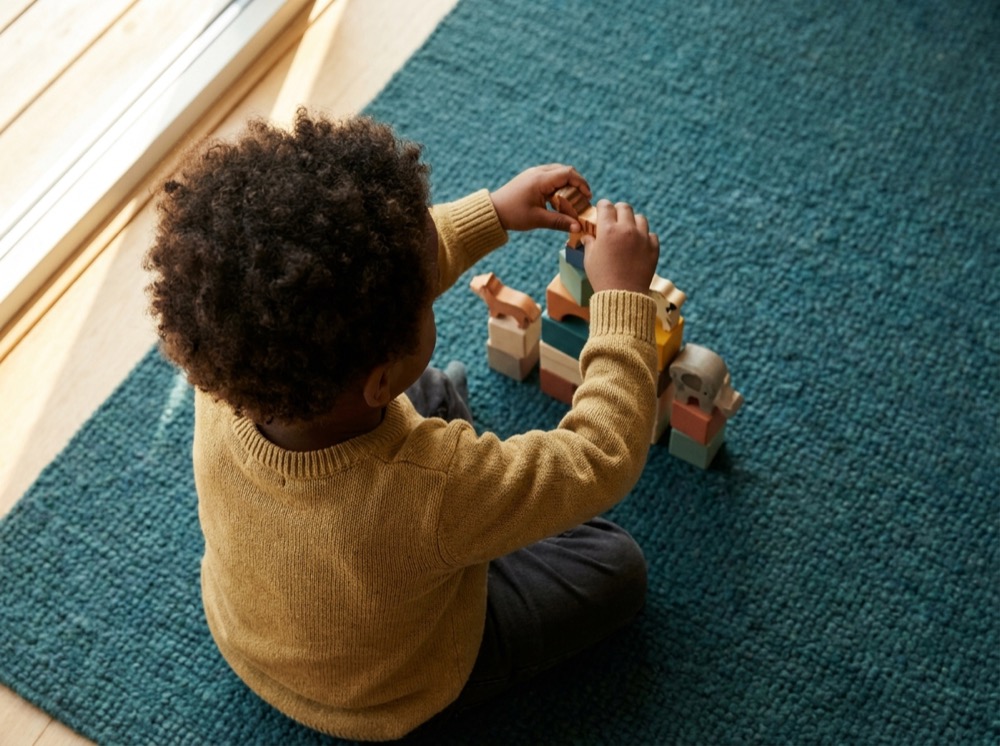 A young child, seen from behind, plays with colourful wooden blocks on a rug.